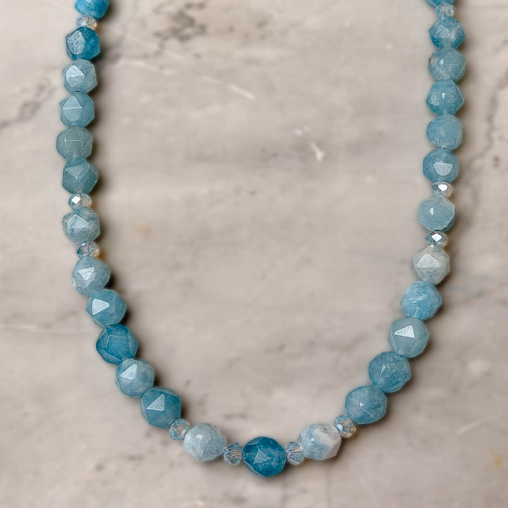 A beaded necklace made of faceted light blue and medium blue Aquamarine beads, with a few small clear Swarovski crystal accents, displayed on a light beige marble surface.