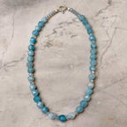 A beaded Aquamarine necklace made of faceted light blue beads with a few translucent Swarovski crystals and a silver clasp, displayed on a light gray marble surface.