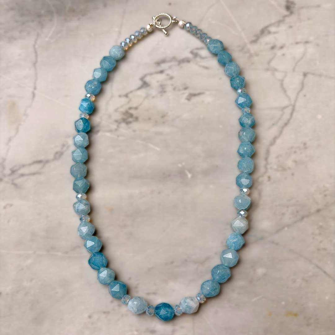 A beaded Aquamarine necklace made of faceted light blue beads with a few translucent Swarovski crystals and a silver clasp, displayed on a light gray marble surface.