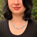 A close-up of a woman wearing a light blue faceted bead necklace, shown from the chest up against a blurred green outdoor background.