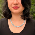 A close-up of a woman wearing a light blue faceted bead necklace, shown from the chest up against a blurred green outdoor background.