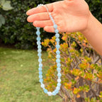 Light blue Aquamarine and sparkling Swarovski crystal bead necklace held up by a person's hand, with a blurred outdoor garden background.