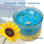 Blue and yellow candle in a jar with decorative elements, adorned with Amazonite, Quartz, Aventurine, Tanzanite, turtle and seashell next to a sunflower, beach in the background.