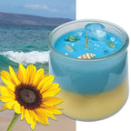 Candle with blue and yellow layers next to a sunflower and beach scene