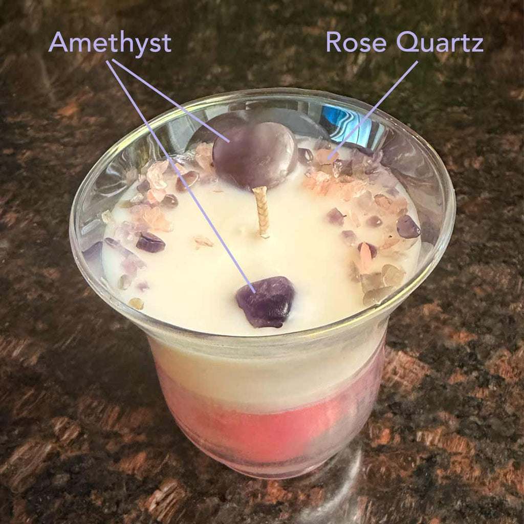 Tulip shaped vessel with white and pink layered wax. Adorned with a Amethyst heart and sprinkles in Amethyst and Rose Quartz. Surrounded by roses and orchids. Callouts define the Amethyst and Rose Quartz. 