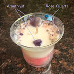 Tulip shaped vessel with white and pink layered wax. Adorned with a Amethyst heart and sprinkles in Amethyst and Rose Quartz. Surrounded by roses and orchids. Callouts define the Amethyst and Rose Quartz. 