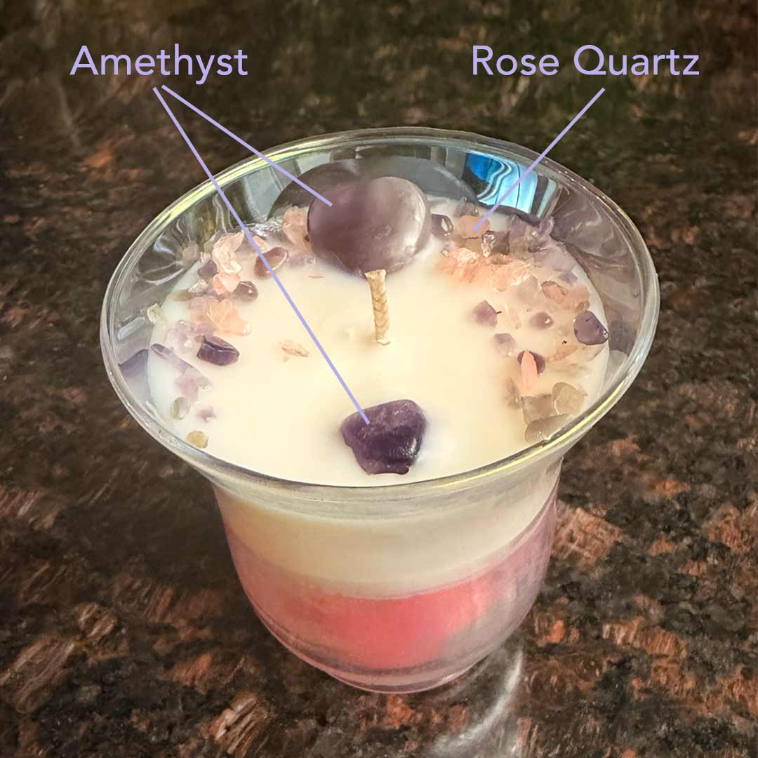 Tulip shaped vessel with white and pink layered wax. Adorned with a Amethyst heart and sprinkles in Amethyst and Rose Quartz. Surrounded by roses and orchids. Callouts define the Amethyst and Rose Quartz. 